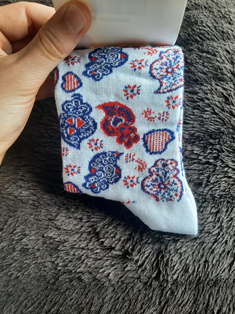 May include: White socks with a repeating pattern of red, white, and blue paisley and floral designs. The socks are held by a hand and are displayed against a dark, textured background.