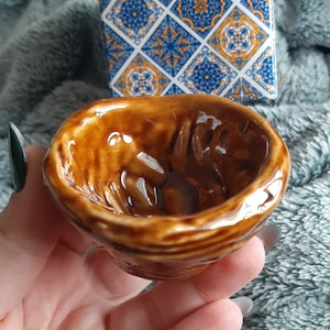 May include: A small, brown ceramic bowl with a glossy finish. The bowl is held in a hand, with a patterned box in the background featuring blue and gold geometric designs. The bowl's interior has a textured surface.