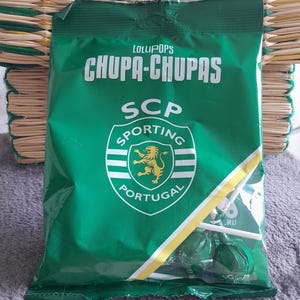Puede incluir: Una bolsa verde y blanca de chupetines Chupa Chups con el logotipo del Sporting Clube de Portugal. La bolsa está sobre una superficie tejida verde y amarilla.