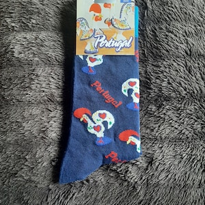 May include: A pair of navy blue socks featuring a repeating pattern of the Rooster of Barcelos, a symbol of Portugal. The socks have a white, red, and blue color scheme. The packaging includes the word "Portugal".