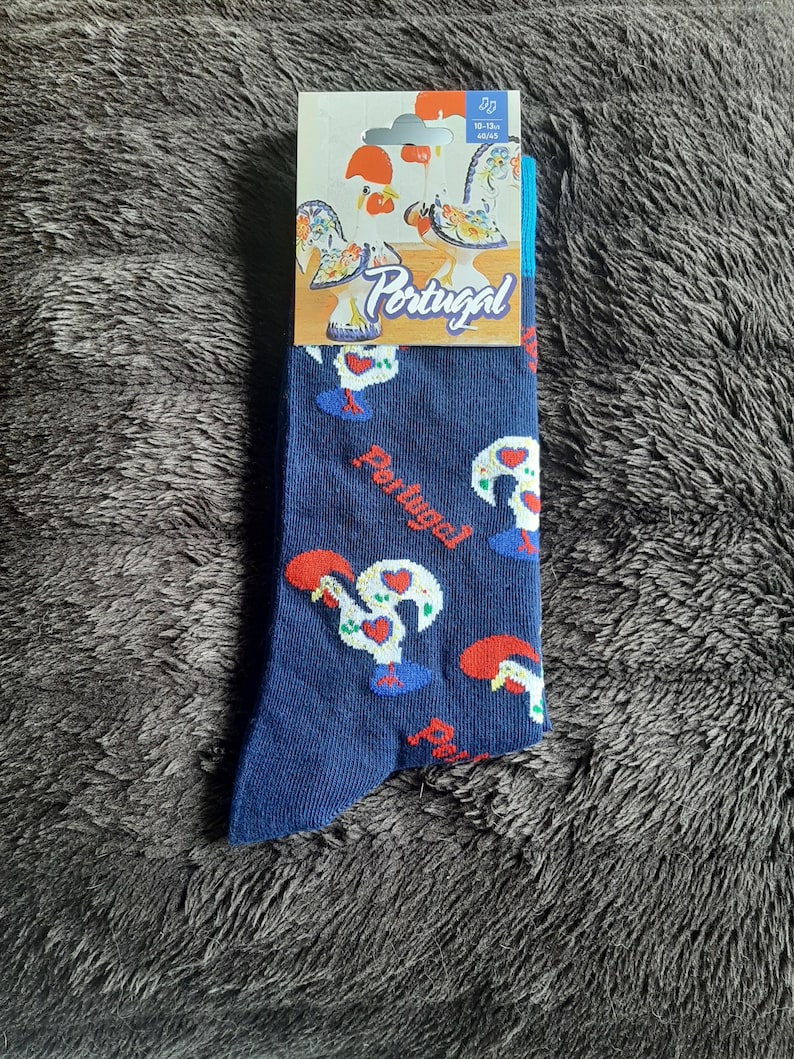 May include: A pair of navy blue socks featuring a repeating pattern of the Rooster of Barcelos, a symbol of Portugal. The socks have a white, red, and blue color scheme. The packaging includes the word "Portugal".