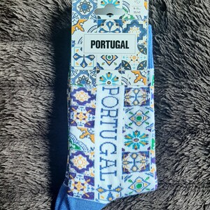 May include: A pair of socks with a Portuguese theme. The socks are white with a blue toe and heel, and feature a repeating pattern of colorful geometric shapes and the word "PORTUGAL". The packaging also displays the word "PORTUGAL".