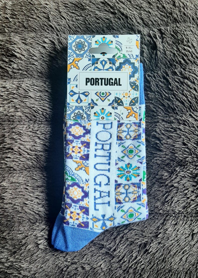 May include: A pair of socks with a Portuguese theme. The socks are white with a blue toe and heel, and feature a repeating pattern of colorful geometric shapes and the word "PORTUGAL". The packaging also displays the word "PORTUGAL".