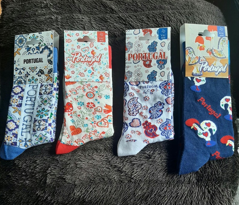 May include: Four pairs of socks with Portuguese-themed designs. The socks feature patterns of flowers, roosters, and traditional tile motifs in red, blue, and white. Each pair is packaged with a cardboard header displaying the word "Portugal".