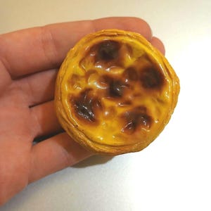 Op de afbeelding: Een enkele pastel de nata, een Portugese custardtaart, met een goudgele custardvulling en een donkere, gekarameliseerde bovenkant. Het gebak heeft een schilferige korst. Het dessert wordt in een hand gehouden tegen een witte achtergrond.