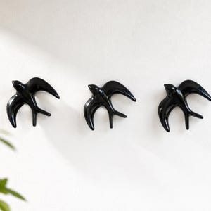 May include: Three glossy black ceramic swallow bird wall decorations arranged in a horizontal row. Each bird is depicted in a flying position with wings spread wide and a forked tail, featuring a smooth, reflective surface and curved, aerodynamic lines.