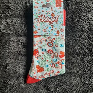 May include: A pair of white socks with a red heel and toe, featuring a floral pattern with red, blue, and green accents. The socks are packaged with a tag that reads "Portugal" and size 9-11.