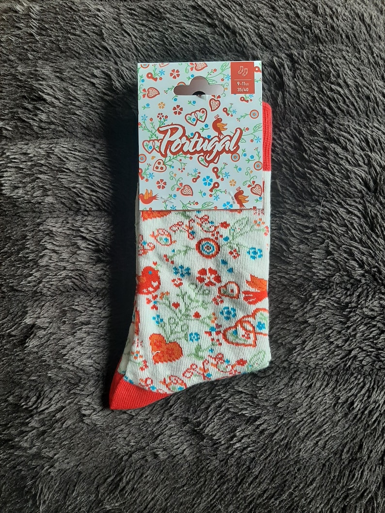 May include: A pair of white socks with a red heel and toe, featuring a floral pattern with red, blue, and green accents. The socks are packaged with a tag that reads "Portugal" and size 9-11.