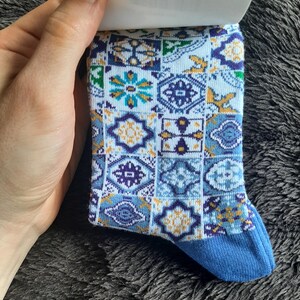 May include: A patterned sock with a blue toe and a design of geometric shapes in shades of blue, white, and yellow. The sock is held by a hand, and the background is a dark, textured surface. The sock's design includes floral and geometric patterns.