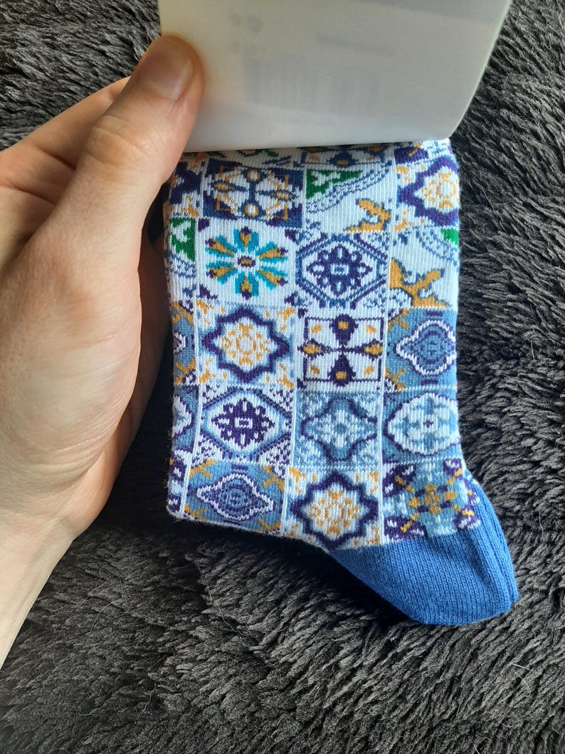 May include: A patterned sock with a blue toe and a design of geometric shapes in shades of blue, white, and yellow. The sock is held by a hand, and the background is a dark, textured surface. The sock's design includes floral and geometric patterns.