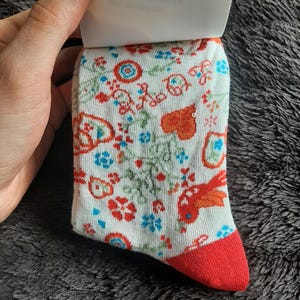 May include: A pair of white socks with a colorful floral pattern and a red toe. The design includes red, orange, and blue flowers and leaves. The socks are held by a hand.