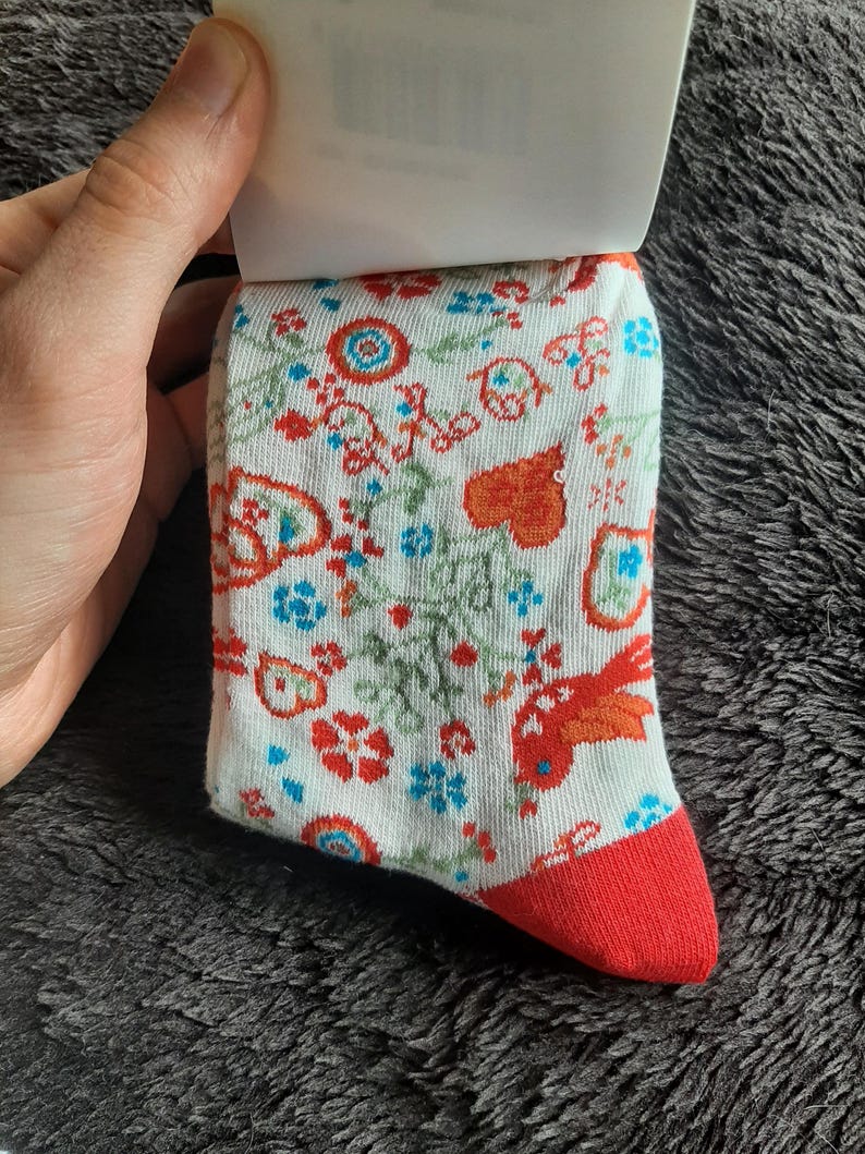 May include: A pair of white socks with a colorful floral pattern and a red toe. The design includes red, orange, and blue flowers and leaves. The socks are held by a hand.