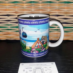 May include: A white ceramic mug featuring a colourful illustration of Madeira, including a cable car, traditional houses with thatched roofs, and flowers. The word Madeira is printed on the inner rim. A white card with black line art is placed in front.