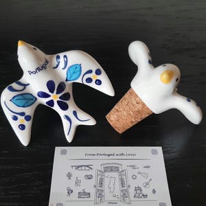 May include: Two ceramic swallow ornaments, one with blue and yellow floral patterns and the word Portugal, the other attached to a cork bottle stopper. A white card features a line drawing of a map and the text From Portugal with Love.