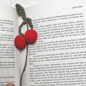 Puede incluir: Marcapáginas de ganchillo hecho a mano con dos cerezas rojas y hojas verdes. El marcapáginas está colocado en un libro con el texto "JANE EYRE" visible. Un accesorio único para los amantes de la lectura.