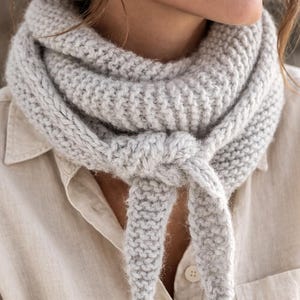 Puede incluir: Bufanda de punto grueso gris claro atada con un nudo. La bufanda está hecha de un hilo suave y texturizado y está envuelta alrededor del cuello. El fondo es una camisa de color claro.