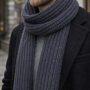 Op de afbeelding: Een donkergrijze, geribbelde gebreide sjaal gedrapeerd om de nek van een persoon. De sjaal is dik en lijkt gemaakt van een zacht, warm materiaal. De persoon draagt een donkere jas en een grijze trui is zichtbaar onder de sjaal.