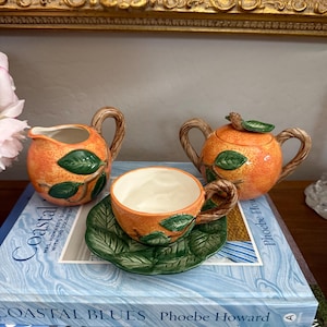 May include: A ceramic tea set with an orange fruit design. The set includes a creamer, a teapot, a teacup, and a saucer. The handles are designed to look like tree branches, and the leaves are green. The set is displayed on top of a stack of books.