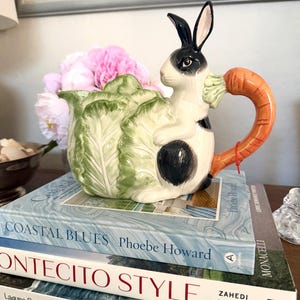 May include: A ceramic teapot featuring a rabbit design with a carrot handle. The teapot is white with black spots and a black and white rabbit head. The teapot is sitting on a stack of books with the titles "Coastal Blues" and "Montecito Style".