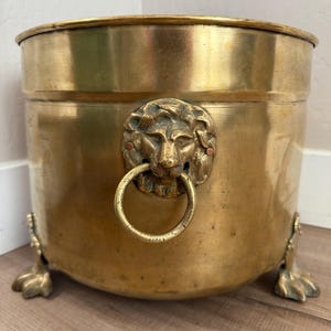 Puede incluir: Una gran jardinera de latón antiguo con un asa de anillo con cabeza de león y patas de garra. La jardinera tiene forma redonda y un acabado pulido. La cabeza de león está detallada con un anillo. Es un artículo decorativo.