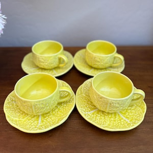 May include: Four matching yellow ceramic teacups and saucers. The cups and saucers have a textured, embossed design. The saucers are shaped like lemon slices. The cups have handles.