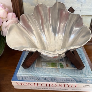 Puede incluir: Un cuenco decorativo plateado en forma de concha con borde festoneado, sostenido por patas de madera oscura. El cuenco se encuentra sobre una pila de libros con los títulos "Coastal Blues" y "Montecito Style".