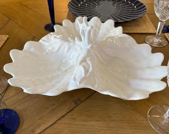 May include: A white, leaf-shaped serving dish with a textured, glossy finish. The dish is placed on a wooden table, alongside blue glassware and a dark blue plate. The dish has a central, raised decorative element.