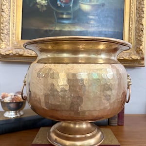 May include: A hammered brass planter with a pedestal base and lion's head ring handles. The planter has a wide, flared rim and a textured surface. It is displayed on a stack of books, with a painting in the background.