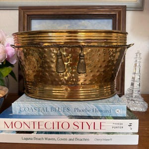 Puede incluir: Una cubitera de latón martillado con dos pequeñas borlas colgando de las asas. La cubitera está colocada encima de tres libros: "Coastal Blues", "Montecito Style" y "Laguna Beach Waves, Coves and Beaches".