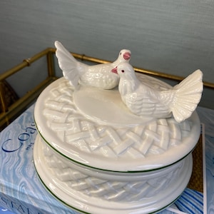 Puede incluir: Una caja de baratijas de cerámica blanca con un diseño de cesta tejida y un borde verde. Dos palomas blancas con detalles rosas están posadas en la tapa. La caja está encima de un libro.