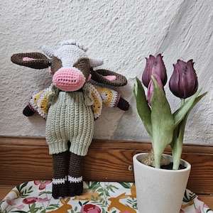 May include: A handmade crochet cow doll with brown and white details, wearing a green jumpsuit and a white hat. The doll is standing next to a small white pot with three dark purple tulips. The doll is on a tray with a floral and rabbit pattern.