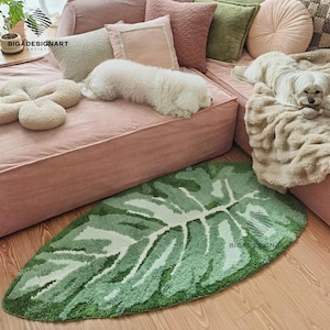 May include: A green leaf-shaped rug with a white pattern, placed on a wooden floor. The rug is surrounded by a pink sofa with various pillows and two small white dogs. The rug has the text "BIGADESIGNART" on it.
