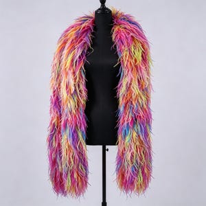 Pode incluir: Uma longa boá de penas multicolorida com um gradiente vibrante de penas cor-de-rosa, laranja, amarelas, azuis e roxas. O acessório está drapeado sobre um torso de manequim preto, exibindo uma aparência densa e texturizada com penas longas.
