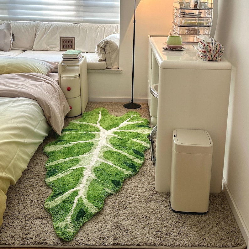 May include: A white and green leaf-shaped rug with a white tree design in the center. The rug is on a light brown carpet. There is a white trash can with a silver lid in the lower right corner of the image.