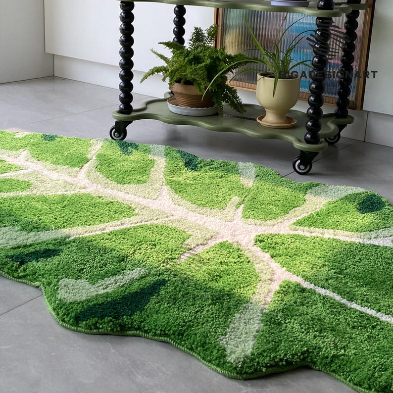 May include: A green and white leaf-shaped bath mat with a fluffy texture. The mat is in front of a green and black rolling cart with plants on the shelves.