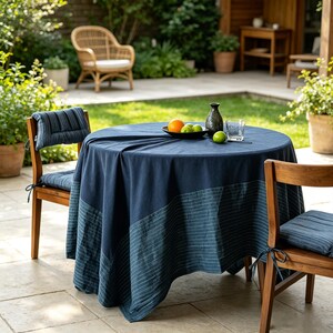 May include: A round table covered with a dark blue tablecloth featuring a horizontal striped border. The table is set with a black bowl containing an orange and green apples, a dark ceramic vase, and a clear glass. Two wooden chairs with blue striped cushions are positioned at the table.