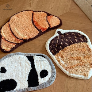 May include: Three decorative rugs in the shape of a croissant, an acorn, and a panda. The croissant rug is brown and orange. The acorn rug is brown, white, and orange. The panda rug is white, black, and gray.