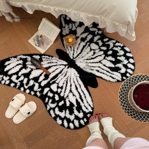 May include: Black and white butterfly-shaped rug with a fluffy texture. The rug is on a wooden floor and there is a book and a candle on the rug.