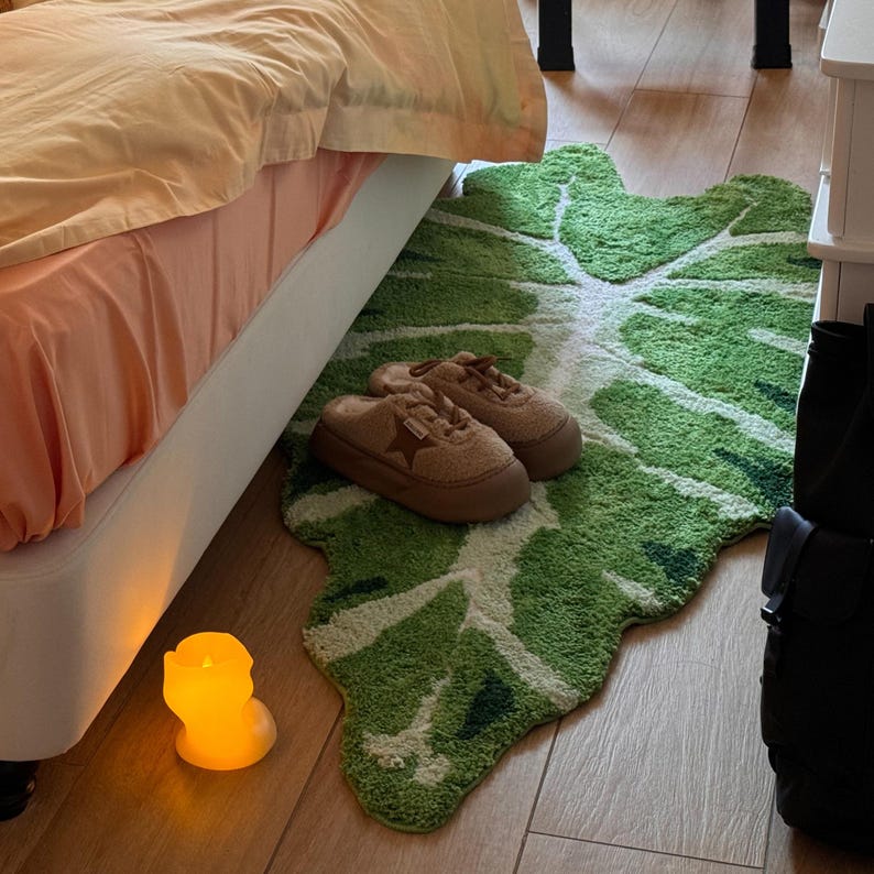 May include: A green leaf-shaped rug with a white outline, placed on a wooden floor. Two brown suede slippers with a star design are on the rug. A lit candle is on the floor next to the rug.