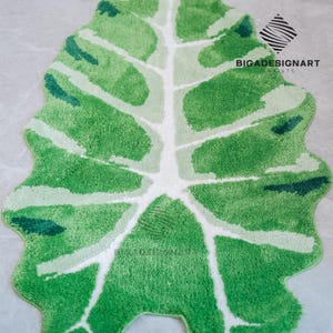 May include: A green and white bath mat shaped like a large leaf. The mat is made of soft, fluffy material and has a white vein pattern.