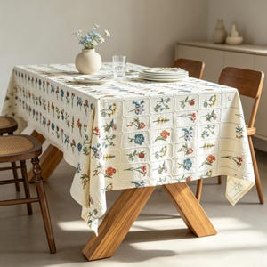 May include: A rectangular cream-coloured tablecloth featuring a repeating grid pattern filled with embroidered colourful wildflowers in shades of blue, orange, red, and green. The fabric has a textured weave and drapes over a wooden table.