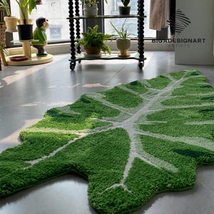 May include: A green and white leaf-shaped rug with a textured surface. The rug is made of a soft, fluffy material and has a realistic leaf design.