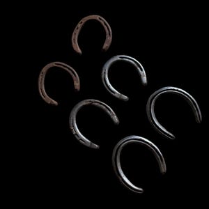 May include: Six horseshoes in various shades of brown and silver are arranged against a black background. The horseshoes are curved, with visible nail holes, suggesting they are used. The arrangement is scattered, with no apparent pattern.