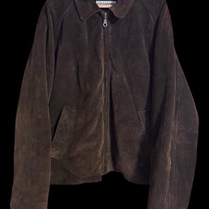 May include: A dark brown suede jacket featuring a classic collar, a full-length front zipper closure with a metal pull, and two side pockets. The interior label displays the text Exposure Urban Wear. The jacket is displayed on a wooden hanger.