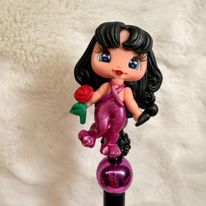 May include: A decorative pen featuring a doll-like figure with black hair, blue eyes, and a red rose. The figure is wearing a magenta outfit and holding a black microphone. The pen has a magenta ball and a black base.