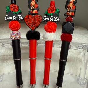 May include: Four black and red pens with different decorative tops. The tops feature a heart-shaped design, a rose, a black and red geometric shape, and a cartoon image of a woman. The pens have the text "Como la Flor" on the top.