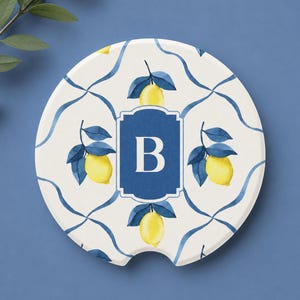Pode incluir: Um porta-copos redondo de cerâmica com fundo branco e um padrão de limões amarelos e folhas azul-escuras ligadas por linhas azuis onduladas. No centro, um emblema retangular azul-escuro com uma borda decorativa exibe a letra B em branco.