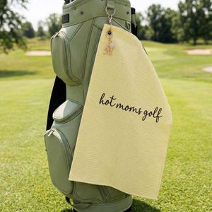K&ouml;nnte beinhalten: Ein hellgelbes Golfhandtuch mit dem Text "hot moms golf" in schwarzer Schrift h&auml;ngt an einer Golftasche. Ein goldener Anh&auml;nger mit dem Buchstaben "M" ist am Handtuch befestigt. Die Golftasche ist hellgr&uuml;n.