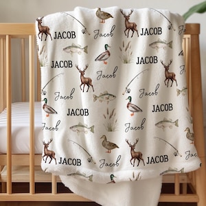 May include: A cream-colored baby blanket with a repeating pattern of deer, fish, ducks, and fishing rods. The name "Jacob" is written in a script font throughout the design. The blanket is draped over a wooden crib.