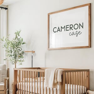 Könnte beinhalten: Ein Kinderbett aus Holz mit einem weißen Laken und einer weißen Decke. Das Bett steht in einem Kinderzimmer mit einer weißen Wand und einem gerahmten Druck, auf dem "CAMERON case" steht.